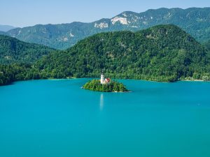 Eine kleine Insel mit Kirche liegt malerisch im blauen Bleder See, im Hintergrund die Julischen Alpen