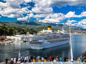 Kreuzfahrtgäste blicken vom Ufer aus auf das majestätische Schiff vor der Kulisse der ligurischen Küste – im Hintergrund erheben sich sanfte Hügel unter klarem Himmel.