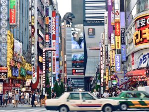 Belebte Straße im Stadtteil Shinjuku in Tokio mit bunten Reklametafeln, Geschäften, Menschen und Autos.