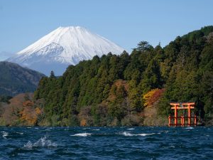 Schneebedeckter Berg Fuji im Hintergrund, grüner Wald mit herbstlichen Farbakzenten und rotes Torii-Tor am Ufer des Ashi-Sees in Hakone
