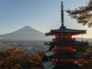 Traditionelle fünfstöckige Chureito-Pagode in Japan mit dem schneebedeckten Berg Fuji im Hintergrund, umgeben von herbstlicher Landschaft und Stadtansicht.