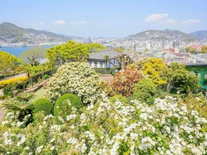 Üppiger Garten mit blühenden Sträuchern und Bäumen im Vordergrund, dahinter Blick auf die Stadt Nagasaki und den Hafen