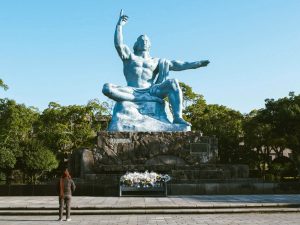Große blaue Friedensstatue auf einem steinernen Sockel im Nagasaki Peace Park, mit erhobenem Arm und üppigem Grün im Hintergrund